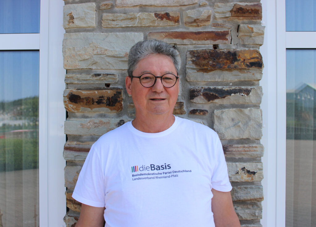 Jörg Gaisbauer - Beisitzer in weißem Basis-T-Shirt vor gemauerter Wand mit weißen Fenstern rechts und link.