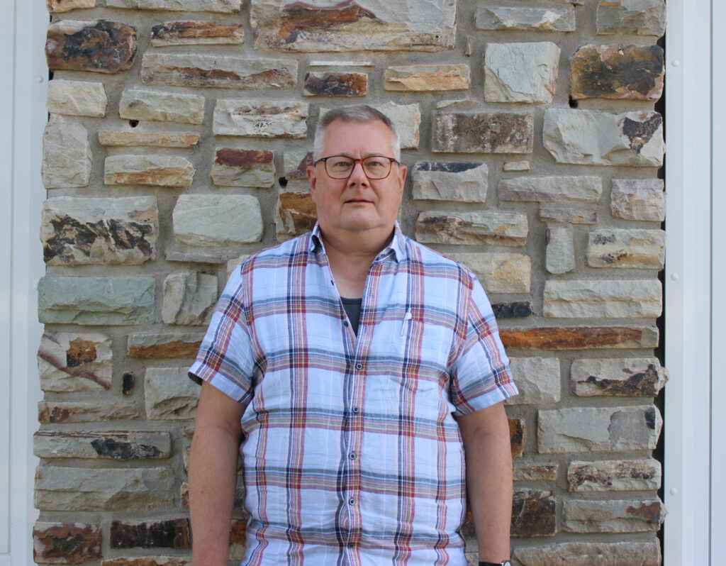 Winfried Sauerborn - Beisitzer im Landesvorstand Rheinland-Pfalz in kariertem Hemd vor einer gemauerter Wand mit weißen Fenstern rechts und links.